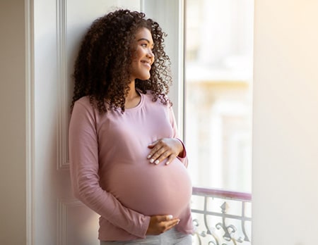 Young bi-racial pregnant woman smiling as she looks out the window and caresses her belly